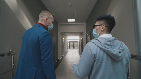 Back view of two men talking and walking in office corridor wear face masks. Covid-19 or Corona Virus Situation in Business Concept. Businessman and his assistant in medical masks walking along hall 库存视频