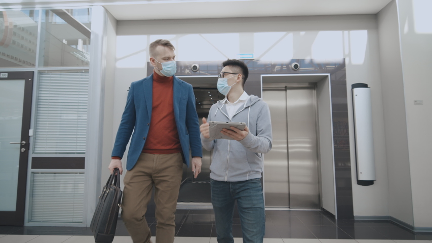 Coronavirus protection. Young employee and boss wearing medical masks walking out of elevator and discussing business over digital tablet. Infection spreading prevention measures Royalty-Free Stock Footage #1048778041