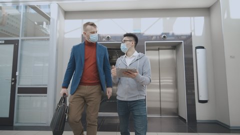 Coronavirus protection. Young employee and boss wearing medical masks walking out of elevator and discussing business over digital tablet. Infection spreading prevention measures