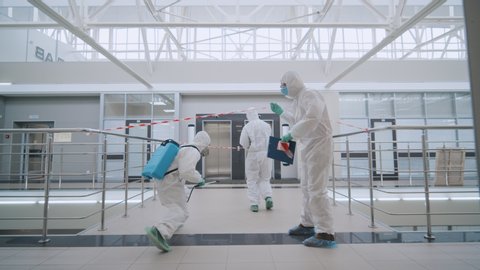 Coronavirus COVID-19 epidemic. Team of scientists in protective coveralls and masks leaving empty business center floor after disinfecting it against biohazard virus.