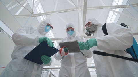 Covid-19 outbreak. Low angle view of disinfectors in mask and protective suit discussing plan of building disinfection using tablet writing report. Scientists treat air from coronavirus infection