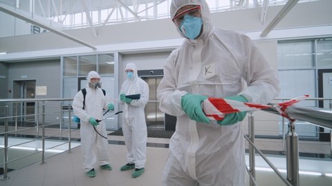 Covid-19 protective measures. Scientists in protective coveralls and masks securing biohazard place with barrier tape before disinfection. Team of workers disinfecting and cleaning business center