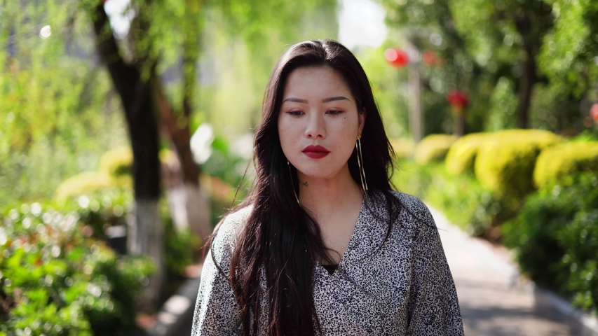 Chinese girl walking through park facing camera. Beautiful young china woman smiling walks outside on sunny day through green nature park towards you.
