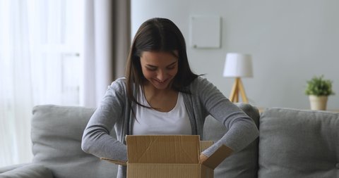 Happy young female client unpacking cardboard parcel, sitting on comfortable sofa at home. Millennial smiling woman unboxing order purchase from internet store, satisfied with fast delivery service