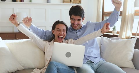 Overjoyed young family couple winners sport fans watching game on laptop celebrating victory goal sit on sofa. Excited man and woman shoppers winning online shopping voucher feeling amazed together.