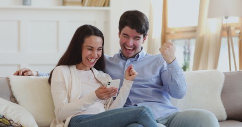 Excited young couple winning giveaway lottery prize in social media app online on smartphone. Happy euphoric millennial man and woman winners celebrating success holding mobile phone sitting on sofa.
