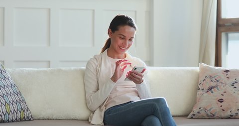 Millennial woman looking at smartphone surfing social media app. Smiling young lady customer texting mobile sms message at home. Happy female customer using smart phone technology sitting on couch