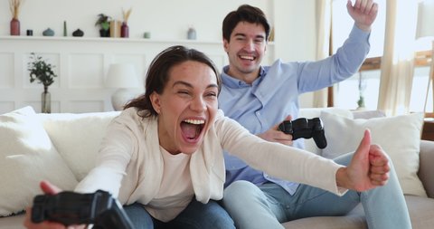 Excited funny young adult couple gamers holding controllers playing video game. Overjoyed man and woman friends winning videogame having fun together on sofa celebrating victory at home.