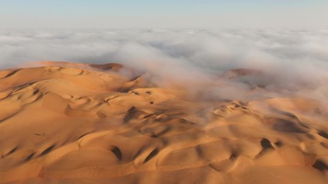 Aerial view of a drone flying over massive sand dunes covered by thick fog clouds at sunrise. Liwa desert, Abu Dhabi, United Arab Emirates.