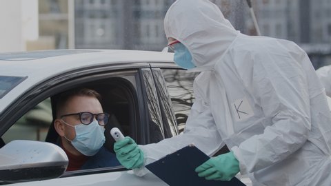 Prevent coronavirus pandemic outbreak. Doctors in hazmat suits checking body temperature of car driver traveller in facial mask and disinfecting his car with detergent spray before letting him drive