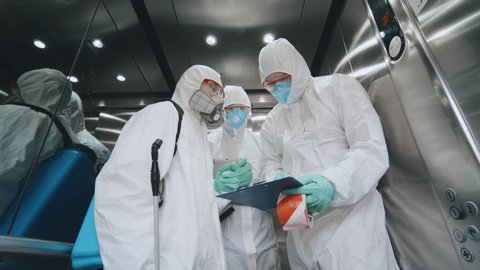 Disinfection from covid-19 coronavirus. Scientist instructing colleagues about disinfection wearing protective suits and masks lifting in elevator. Team of disinfectors working in office building