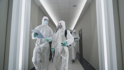 Epidemic of coronavirus. Team of three disinfectors in protective masks and coveralls walking along empty office corridor after successful disinfection from coronavirus bacteria. Covid-19