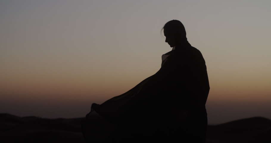 In the Dubai desert at sunset, girl in the wind develops traditional Abaya dress and hijab. 4K Slow Motion Royalty-Free Stock Footage #1048799794