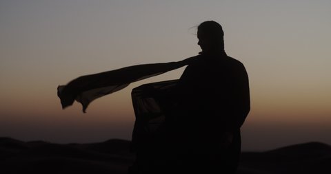 In the Dubai desert at sunset, girl in the wind develops traditional Abaya dress and hijab. 4K Slow Motion