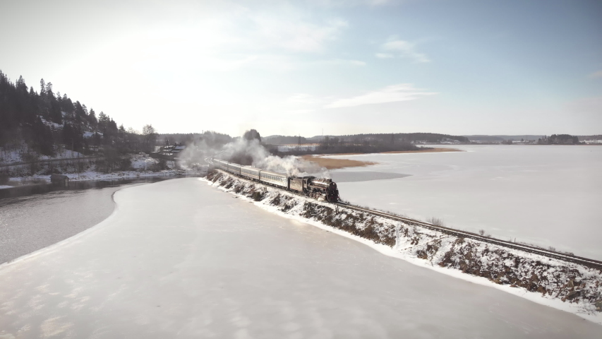 Old train aerial view of steam train running on the tracks across the lake in Karelia. Heritage historic steam locomotive with white smoke shot from above by drone. 