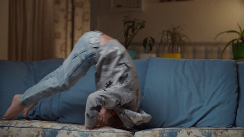 Kid in jeans making somersault in living room. Child making acrobatic movements on couch. Blonde boy fooling around on cozy sofa at evening room.