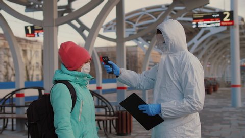 Controlling people's temperature and health at platform of railway station. Medical worker epidemiologist in protective suit screening passenger at public places to check the Covid-19 symptoms