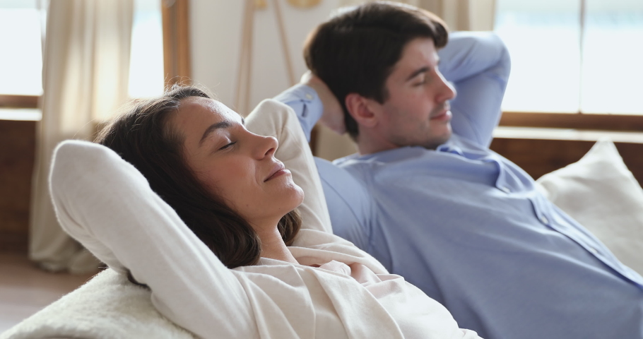 Calm serene young couple relaxing sitting on sofa at home with eyes shut. Smiling lazy husband and wife breathing fresh air, enjoying comfort, no stress in cozy living room holding hands behind head. Royalty-Free Stock Footage #1048820509