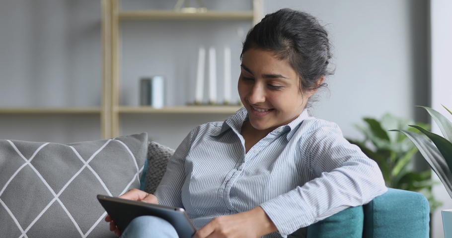 Happy indian millennial girl sitting on comfy sofa, using digital tablet, satisfied with remote work results. Smiling female hindu customer shopping online in internet store, web surfing information. Royalty-Free Stock Footage #1048820551