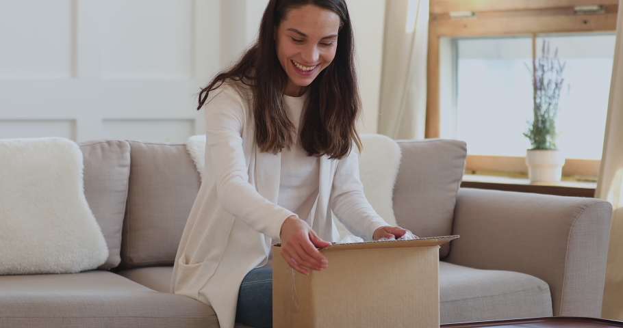 Excited young woman customer opening parcel box at home. Amazed happy girl shopper unboxing fashion purchase sitting on couch. Satisfied female consumer unpacking postal shipping delivery concept Royalty-Free Stock Footage #1048820560