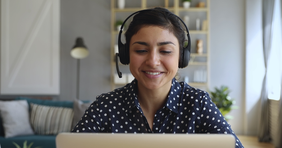 Front view head shot close up smiling indian young girl wearing headset with microphone, looking at web camera on computer, studying online. Motivated hindu tutor giving video call educational class.