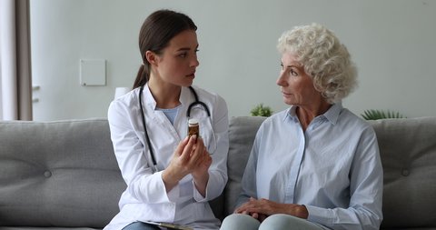 Focused young female therapist sitting on couch with old retired patient, explaining medicine prescription. Serious doctor holding glass bottle with pills, reading side effects to elderly woman.