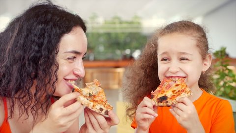 Happy Smiling family of two eagerly eat delicious pizza, Cute little girl daughter with sits with her mother at the table eating italian pizza, looking at each other smiling in response.