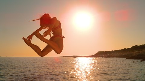 SLOW MOTION, LENS FLARE, COPY SPACE: Cinematic shot of an agile woman jumping off a cliff and into the ocean at sunset. Golden sunbeams shine on energetic woman diving into the refreshing Adriatic sea