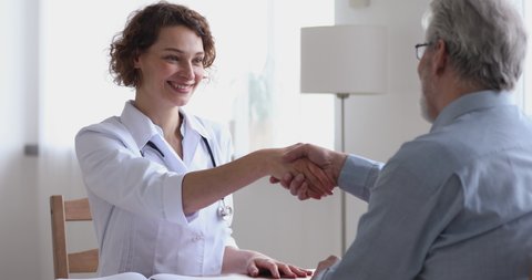 Smiling friendly female physician giving healthcare advice handshaking senior adult male patient. Happy doctor and older client shake hands at medical office expressing respect, gratitude and trust.