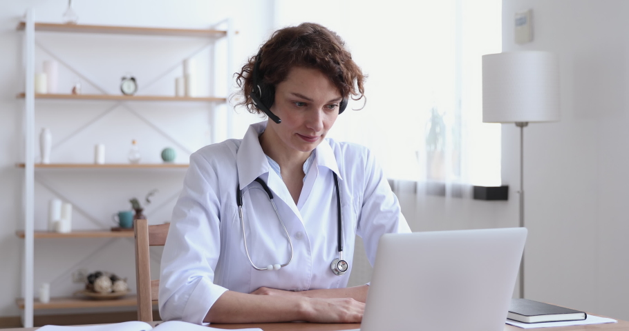 Female medical assistant wears white coat, headset video calling distant patient on laptop. Doctor talking to client using virtual chat computer app. Telemedicine, remote healthcare services concept. | Shutterstock HD Video #1048839271