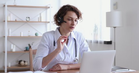 Female medical assistant wears white coat, headset video calling distant patient on laptop. Doctor talking to client using virtual chat computer app. Telemedicine, remote healthcare services concept.