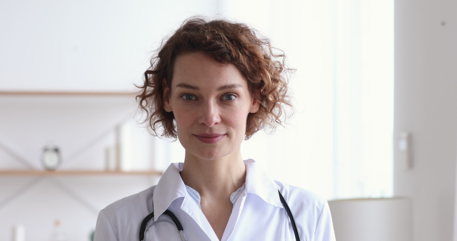 Happy young european woman doctor wearing white medical coat and stethoscope looking at camera. Smiling female physician posing in hospital office. Positive general practitioner close up face portrait | Shutterstock HD Video #1048839322