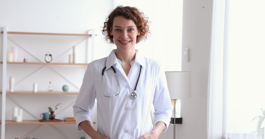 Confident millennial female therapist posing standing in medical office. Smiling young woman professional doctor looking at camera. Portrait of happy physician, general practitioner wears white coat. | Shutterstock HD Video #1048839325