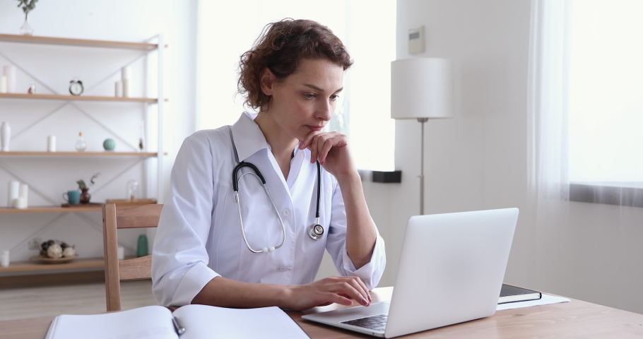 Serious young woman doctor physician wearing white coat using laptop computer writing notes at workplace. Female professional medic consulting patient distantly in online chat sit at desk in hospital. Royalty-Free Stock Footage #1048839328
