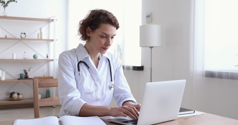 Serious young woman doctor physician wearing white coat using laptop computer writing notes at workplace. Female professional medic consulting patient distantly in online chat sit at desk in hospital.