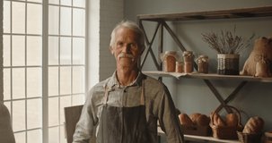 Expressive elderly caucasian moustached man working as a experienced baker at traditional bakery shop, looking at camera and joyfuly smiling portrait shot 4k footage
