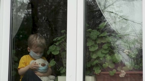 Bored child on home quarantine. Boy in protective medical mask wears mask to teddy bear, sits on windowsill and looks out window. Allergy, covid-19 coronavirus pandemic, prevention epidemic.