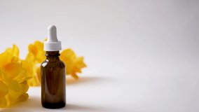 Dark glass bottle with hyaluronic acid or cosmetic serum and yellow spring flowers on white background with copy space. Drops of essential oil falling into container from cosmetic pipette. 4k video. 