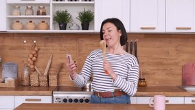 Happy woman dancing and singing in kitchen using her phone with karaoke and listening to music at home. Removes live streaming for social networks.