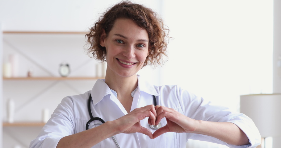 Smiling young woman doctor cardiologist wearing white medical coat and stethoscope showing hands heart shape looking at camera. Cardiology healthcare, love and medicine charity concept, portrait.