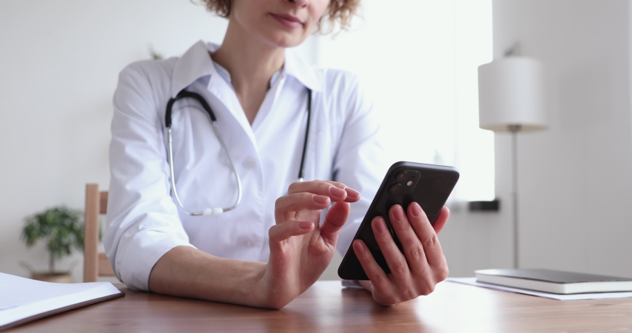 Female doctor wearing white coat holding cell using smart phone apps texting message to remote patient. Practice of medicine and public health care supported by mobile devices concept. Close up view