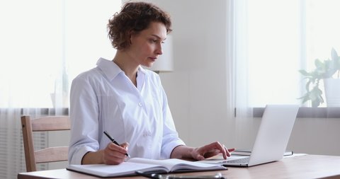Young female nurse writing notes using laptop at workplace desk. Woman professional doctor wearing white coat working on computer checking appointments, doing research or report in hospital office.