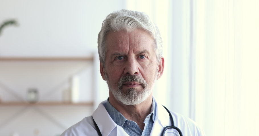 Confident professional senior male doctor wearing white uniform, stethoscope looking at camera. Bearded 60s successful older man physician or gp with dental smile posing for close up face portrait. Royalty-Free Stock Footage #1048870834