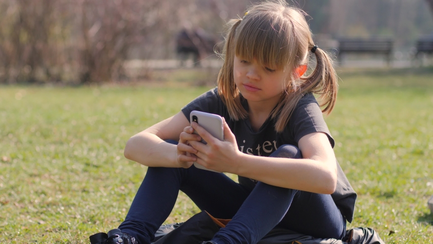Lazy inactive kid girl with smartphone sit and lie on park lawn grass watching device diplay playing game application