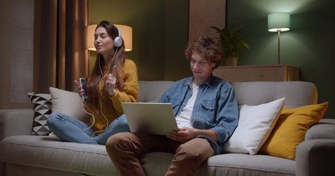 Charming young girl blinking to her boyfriend with laptop computer while she listening to music and dancing in headphones and with smartphone on sofa at home at night.