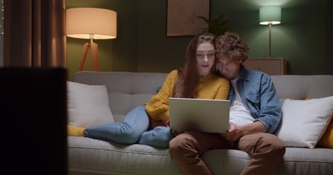 Romantic and happy young couple sitting on the couch in hugs and watching something on the laptop computer in the evening. Indoor.
