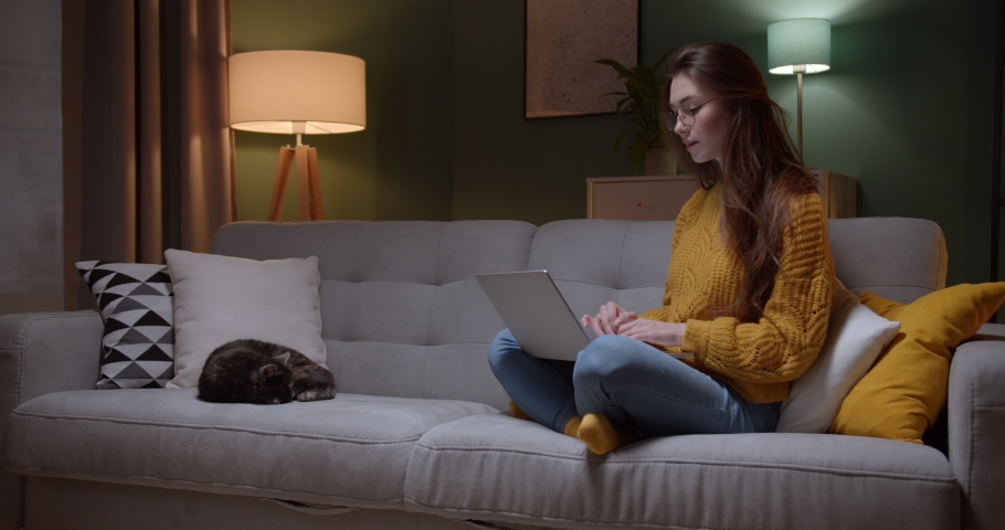 Young good looking Caucasian girl in glasses typing and working on the laptop computer as freelancer while sittng at home in the cozy dark living room with a cat. At night. Stay home | Shutterstock HD Video #1048878394