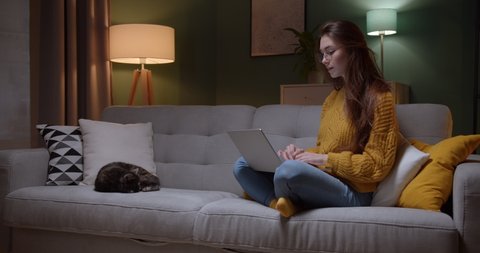 Young good looking Caucasian girl in glasses typing and working on the laptop computer as freelancer while sittng at home in the cozy dark living room with a cat. At night. Stay home