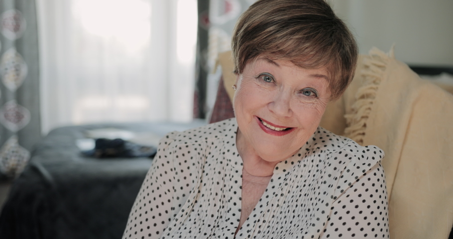 Close up view of cheerful female pensioner sitting on chair at her apartment. Portrait ofgood looking old lady smiling while looking to camera. Concept of peole and positive emotions
