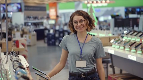 Positive female seller or shop assistant portrait in supermarket store. Woman in blue shirt and empty badge looking at the camera and smiling. Electronic devices on the background. Slow motion